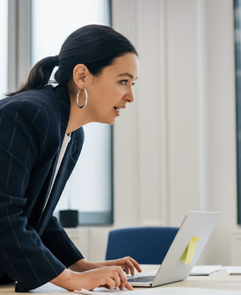 Woman with hoop earrings leaning forward and typing on a laptop at a desk in a bright office.