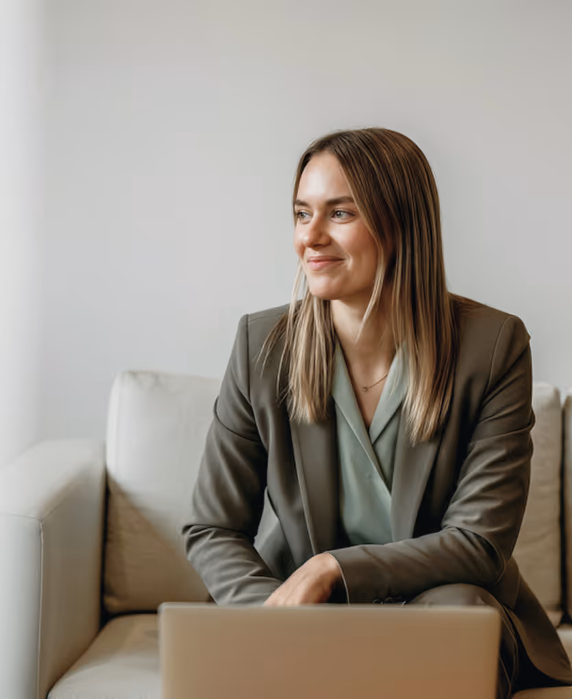 Smiling woman in business attire sitting on a sofa with a laptop in front of her, looking to the side.
