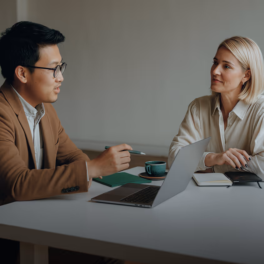 Man and woman have a discussion at a table with laptop, notebooks, and coffee cup.