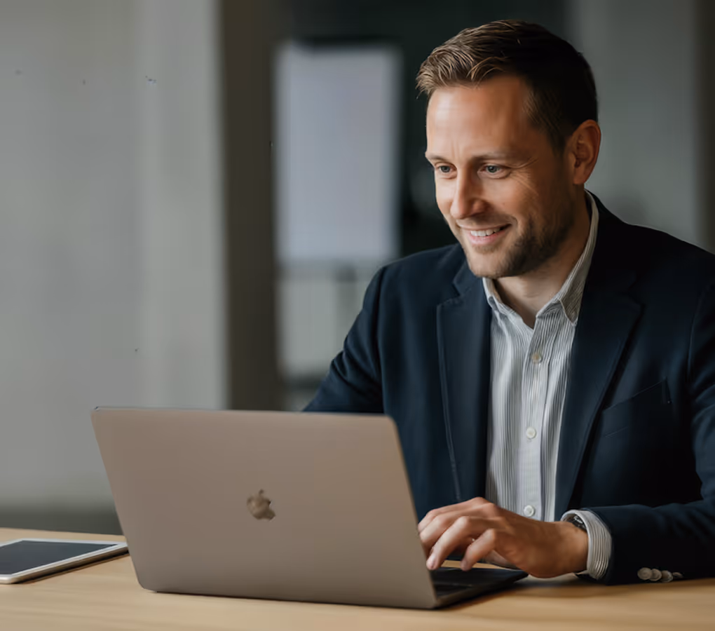 Smiling man in a blazer typing on a laptop at a wooden desk with a tablet nearby.