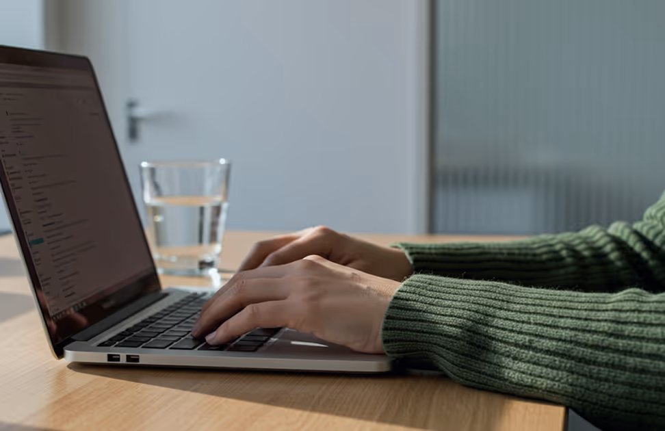 Person typing on a laptop keyboard with a glass of water on a wooden table nearby.