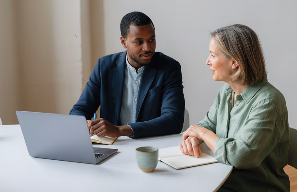 Two people seated at a table discussing, with a laptop, notebook, and coffee mug in front of them.