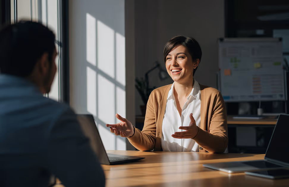 Smiling woman in a brown cardigan talking to a man across a table with laptops in an office.