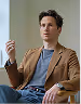 Man with short dark hair wearing a brown blazer and gray shirt sitting on a chair against a white background.
