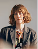 Woman with wavy light brown hair wearing a dark blazer and colorful necklace against a gray background.