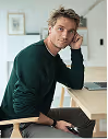 Man in green sweater sitting at wooden desk with laptop, looking at camera.