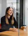 Smiling woman sitting at a table with a takeaway coffee cup in a bright indoor setting.