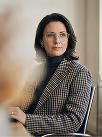 Woman with glasses wearing a brown checkered jacket and black turtleneck, seated by a window.