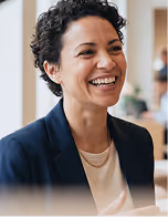 Smiling woman with short curly hair wearing a navy blazer and light shirt indoors.