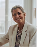 Smiling mature woman with gray hair wearing a beige blazer sitting near a window.