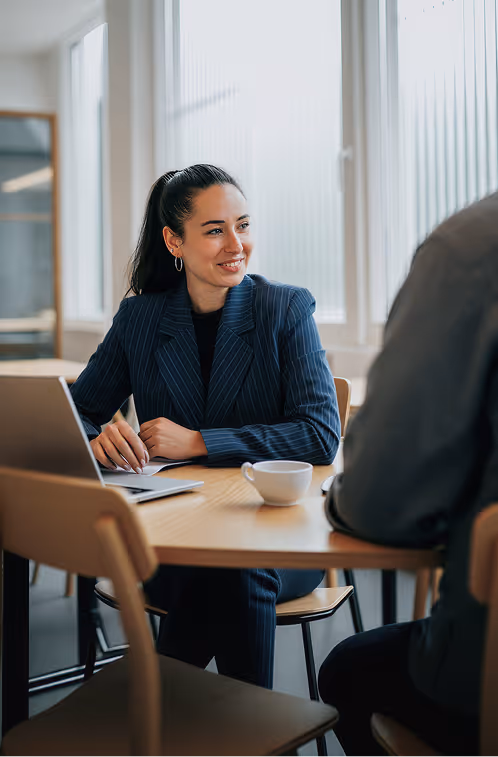 Woman in pinstripe blazer smiling and talking during a meeting at a table with a laptop and coffee cup.