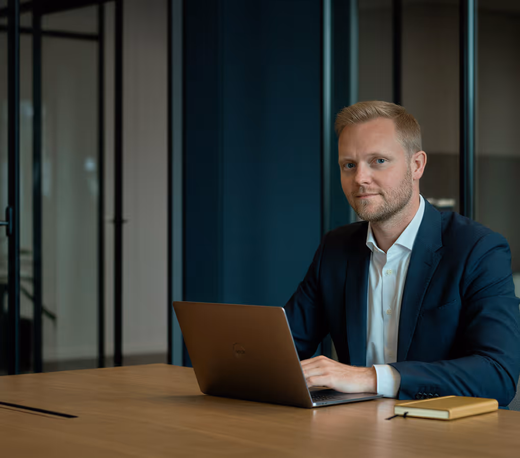 Man in a suit sitting at a desk using a laptop with a notebook nearby in a modern office.