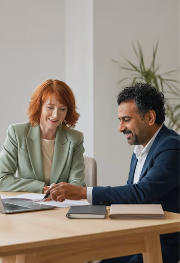 Two people sitting at a table, reviewing documents and using a laptop in a modern office.