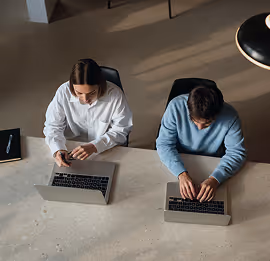 Two people typing on laptops at a table viewed from above.