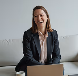 Smiling woman in a black blazer sitting on a couch with a laptop and coffee cup nearby.