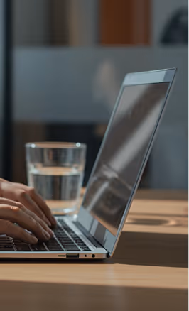 Person typing on a laptop keyboard with a glass of water on a wooden desk.