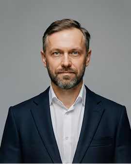 Middle-aged man with beard wearing a dark suit and white shirt against a gray background.