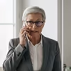 Mature woman with glasses in gray blazer standing in a bright room, looking thoughtful.