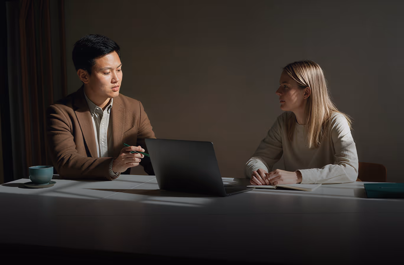 Two people sitting at a table discussing with a laptop and notebook under dim lighting.