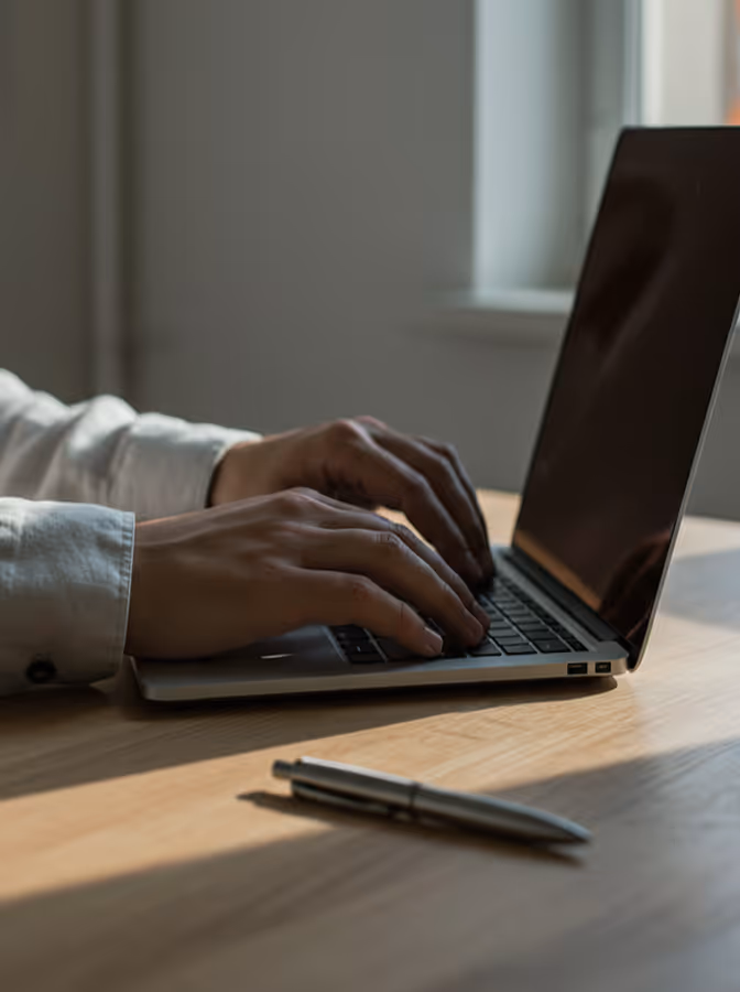 Person typing on a laptop keyboard on a wooden desk with a pen nearby.