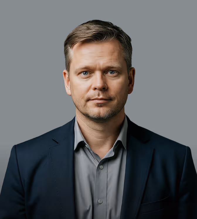 Professional man with short hair and blue eyes wearing a gray shirt and dark blazer on dark background.