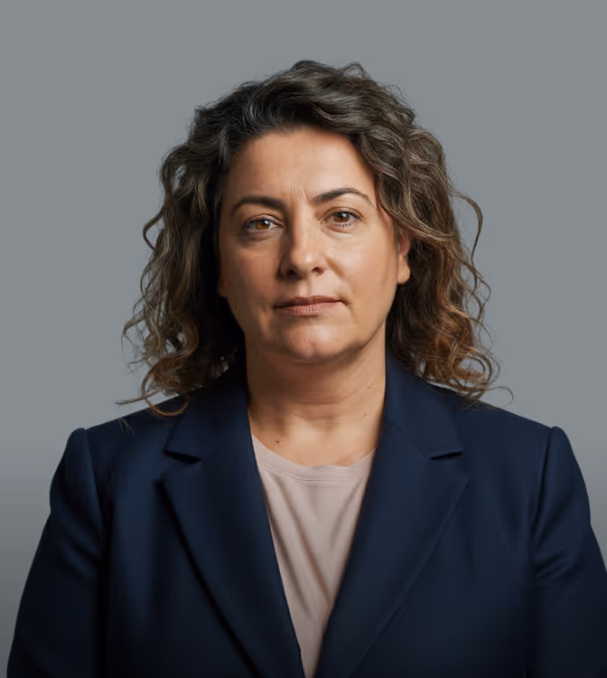 Serious middle-aged woman with curly hair wearing a navy blazer and light shirt on dark background.